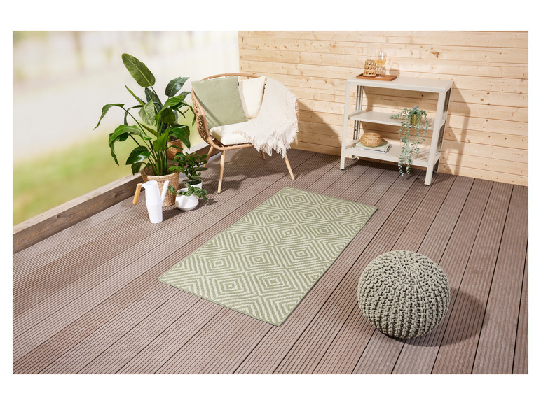 Ein grüner Teppich mit Diamantmuster auf einer Holzterrasse mit einem grauen, gestrickten Hocker.