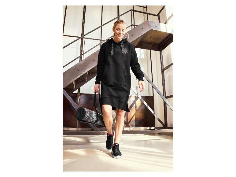 Frau in einem schwarzen Hoodie-Kleid und Puma Sneakern mit einer Yogamatte.
