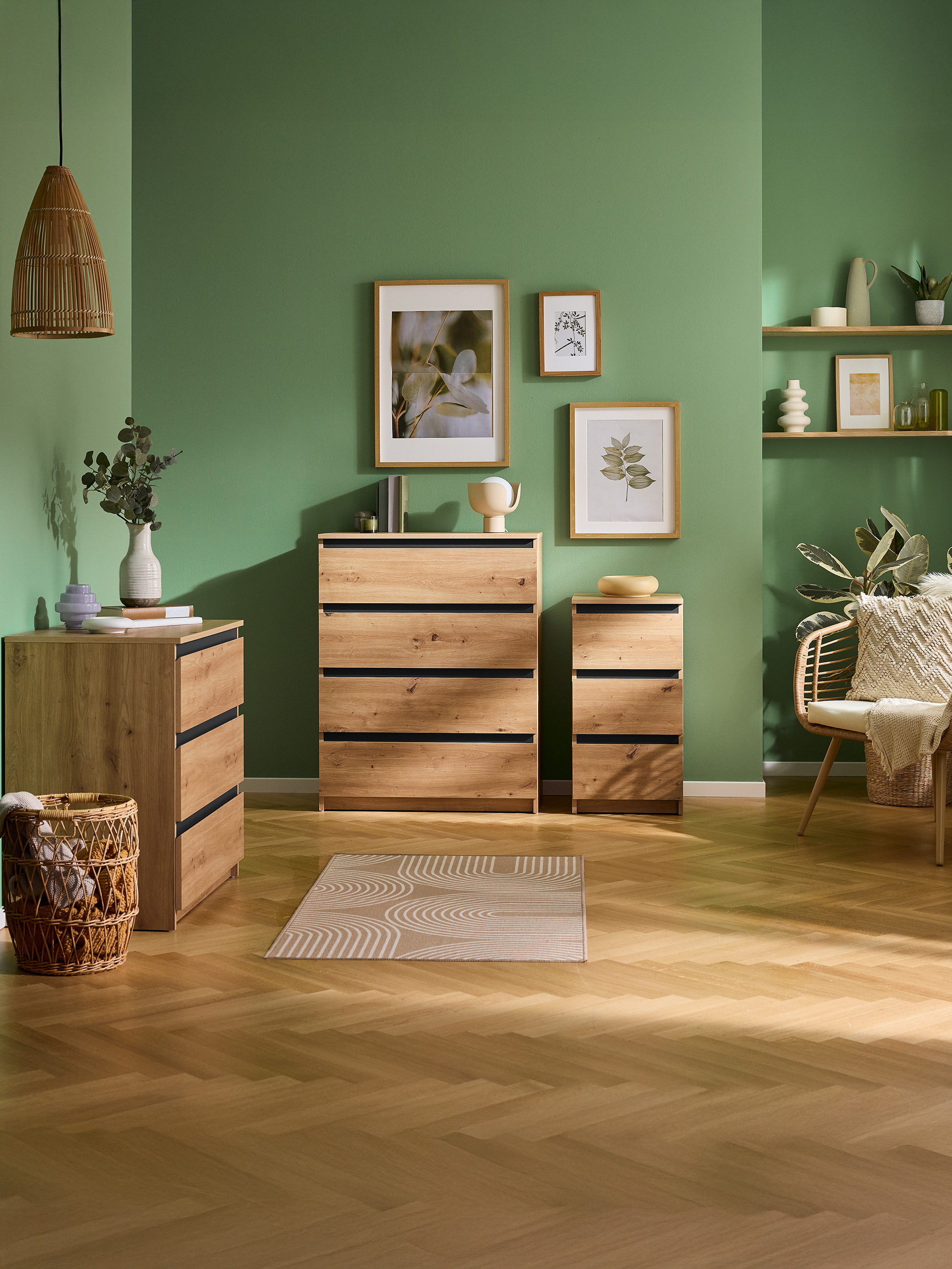 Grünes Zimmer mit Holzkommoden, Regalen und einem Korbstuhl, mit botanischer Kunst.