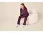 Mädchen in dunkelviolettem Velours-Set mit Kapuzenpullover und Hose, sitzt auf einem weißen Sessel.