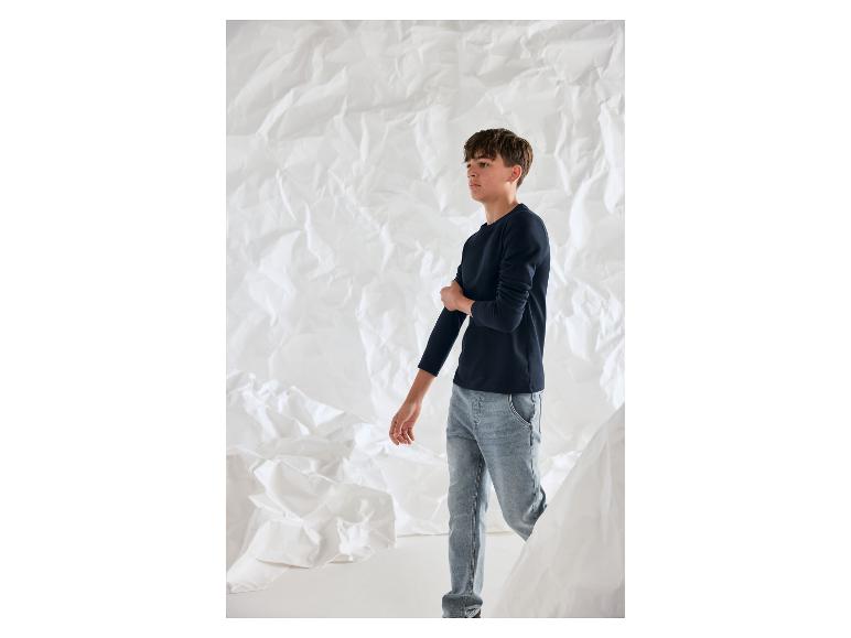 Ein Junge in einem dunkelblauen Langarmshirt und hellen Jeans vor einem zerknitterten weißen Hintergrund.