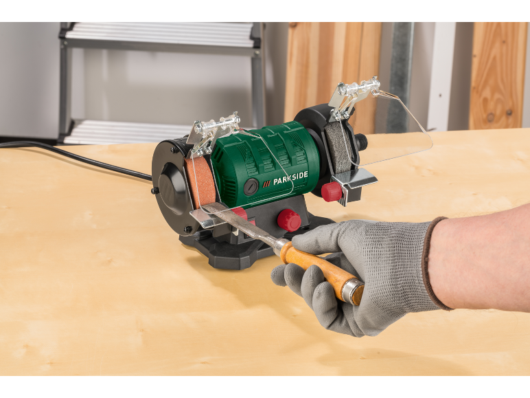 Grüner Doppelschleifbock mit einem Holzmeißel, der von einer behandschuhten Hand geschliffen wird.