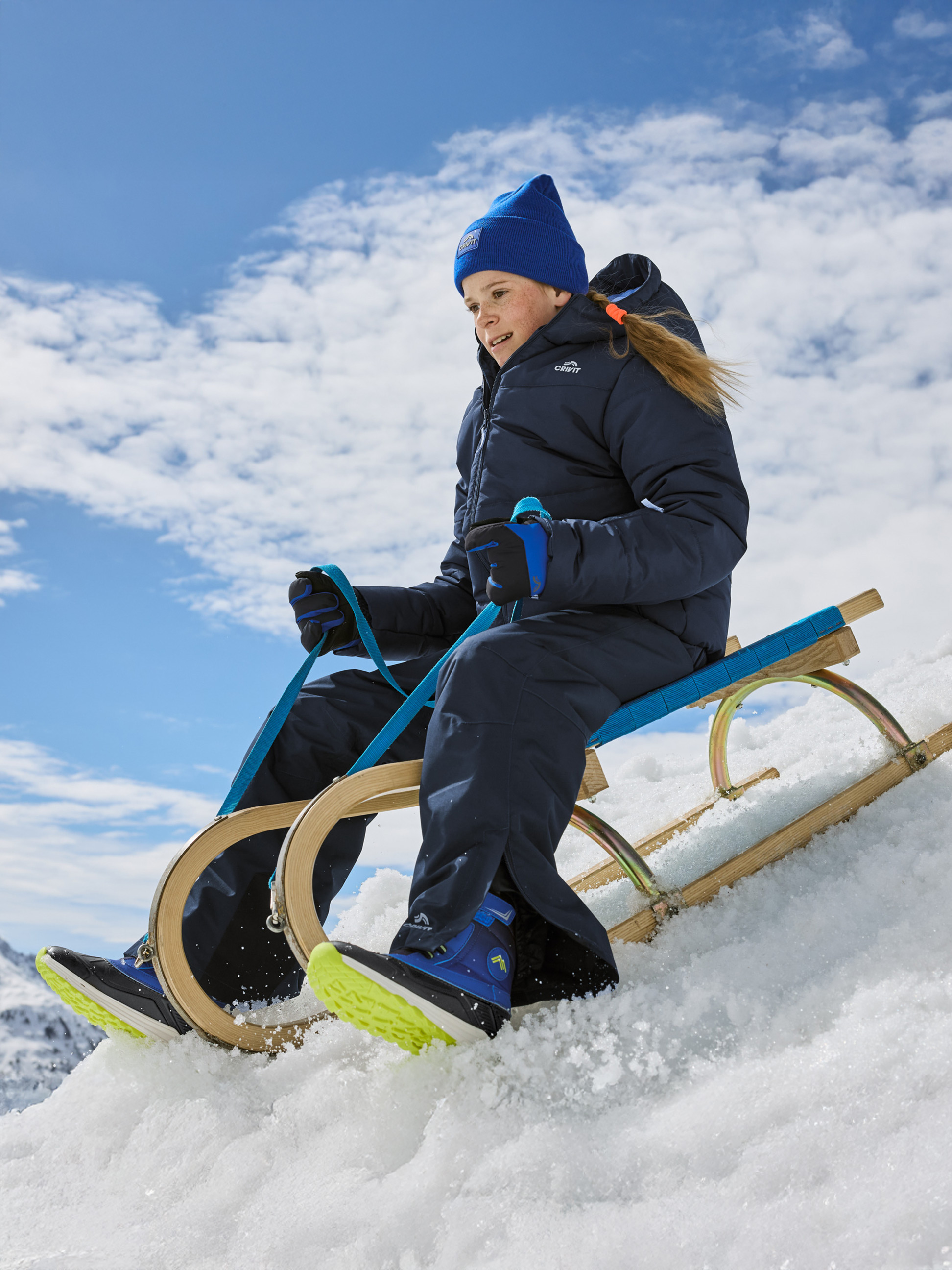 Mädchen in blauem Schneeanzug und Mütze rodelnd im Schnee.