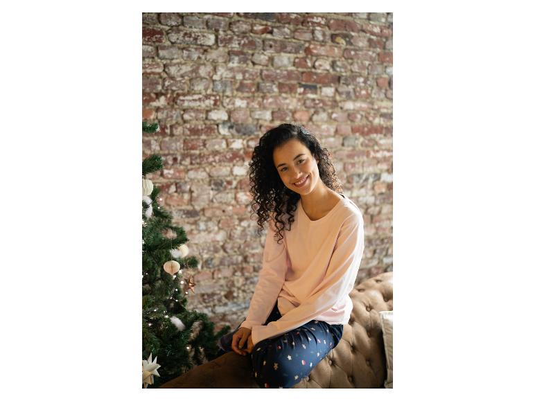 Eine Frau trägt einen rosa Pullover und eine Pyjamahose mit Weihnachtsmotiv und sitzt auf einem Sofa mit einem Weihnachtsbaum daneben.