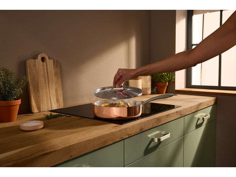 Kochen in einer Kupferpfanne auf Induktionskochfeld, mit Schneidebrett und Kräutern im Hintergrund.