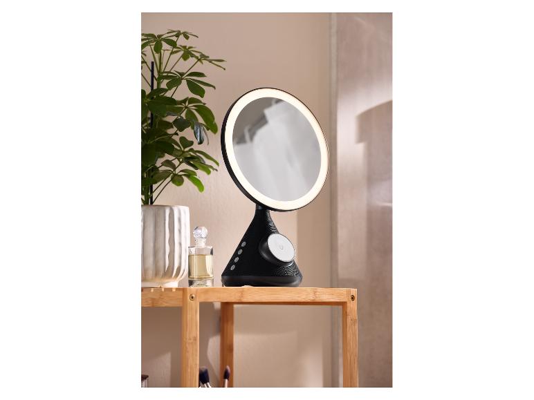 Beleuchteter Kosmetikspiegel mit Bluetooth-Lautsprecher auf Holzregal mit Pflanze und Parfüm.