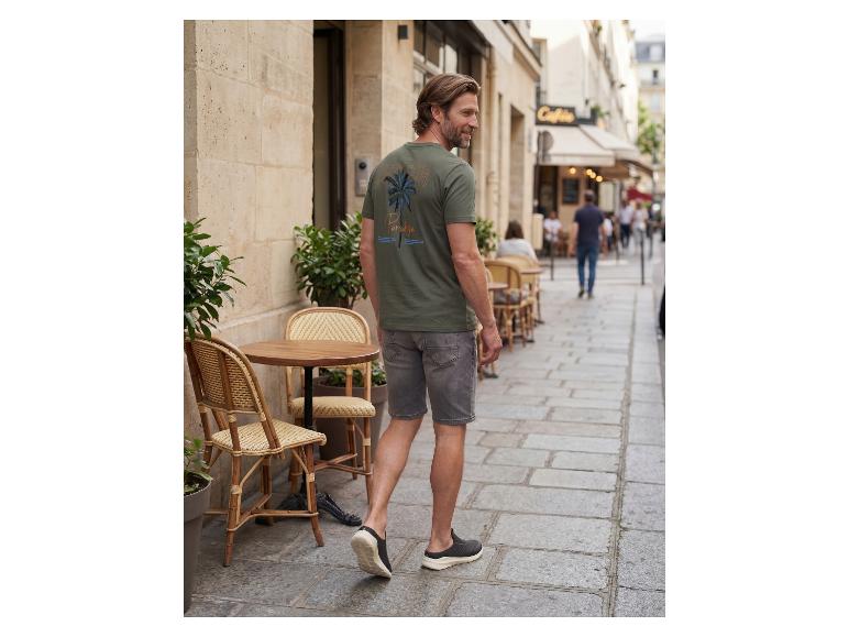 Mann in grünem T-Shirt mit Palmenaufdruck und grauen Shorts geht eine Straße entlang.