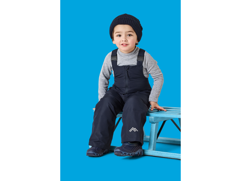 Kind in Skioverall und Strickmütze, sitzt auf einem Holzschlitten.