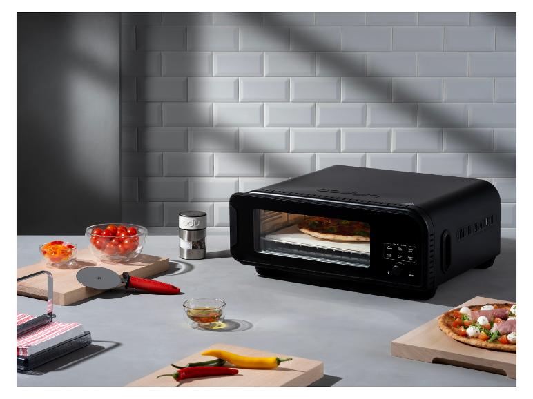 Ein schwarzer Pizzaofen mit einer Pizza darin, umgeben von frischen Zutaten und Küchenutensilien.