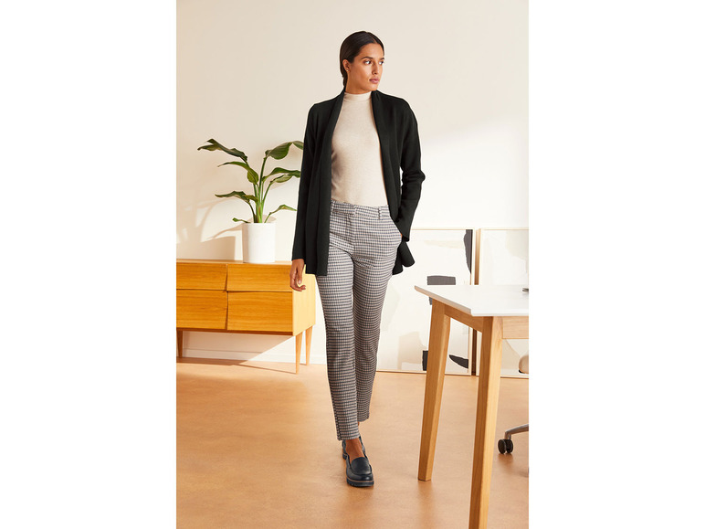 Eine Frau in einem schwarzen Blazer, einem beigefarbenen Pullover und einer karierten Hose.