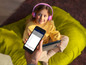 Eine Hand hält ein Telefon mit einer App zum Einstellen von Lautstärkebegrenzung und Spielzeit, im Hintergrund ein Mädchen mit Tablet und Kopfhörern.