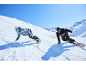 Zwei Snowboarder fahren einen verschneiten Hang unter blauem Himmel hinab.