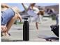 Schwarze Isolierflasche auf einer Betonstufe mit Personen und Skateboard im Hintergrund.