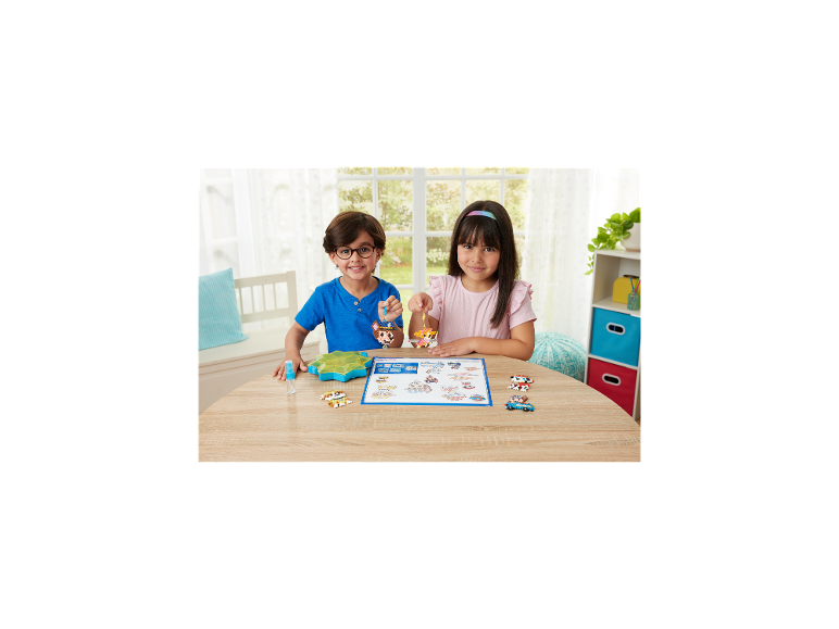 Zwei lächelnde Kinder halten bunte Perlenkreationen an einem Holztisch.