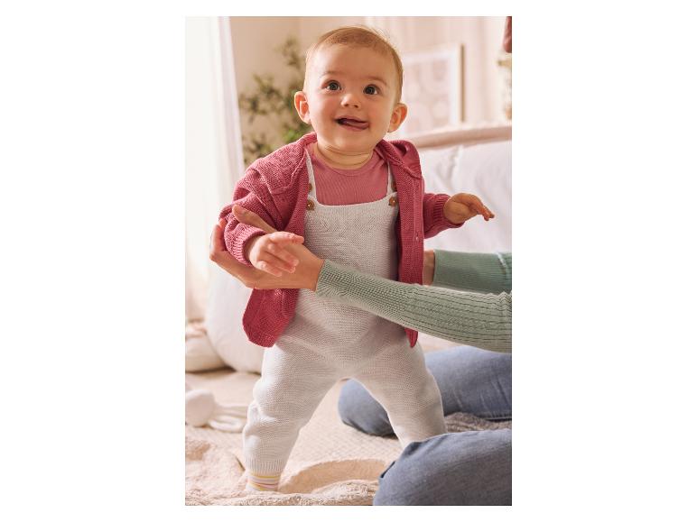 Baby in rosa Strickjacke und weißer Latzhose lernt mit Hilfe eines Erwachsenen laufen.