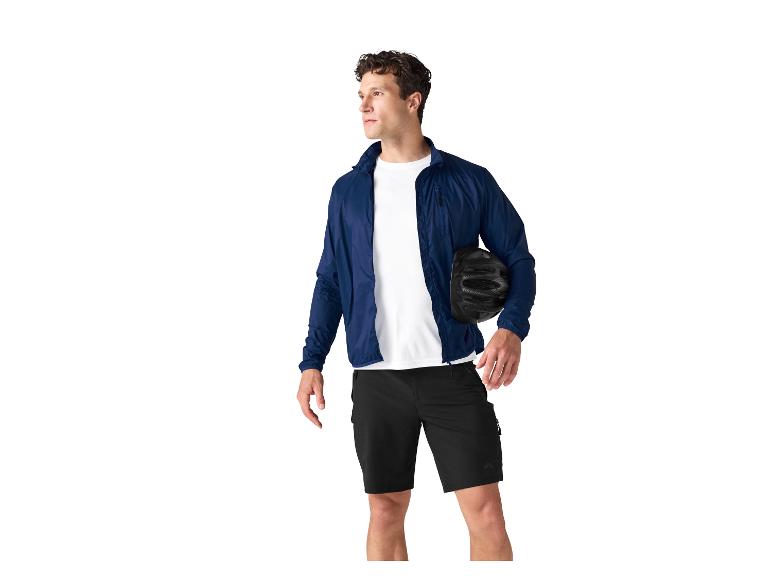 Mann in blauer Jacke, weißem T-Shirt und schwarzen Shorts hält einen Fahrradhelm.