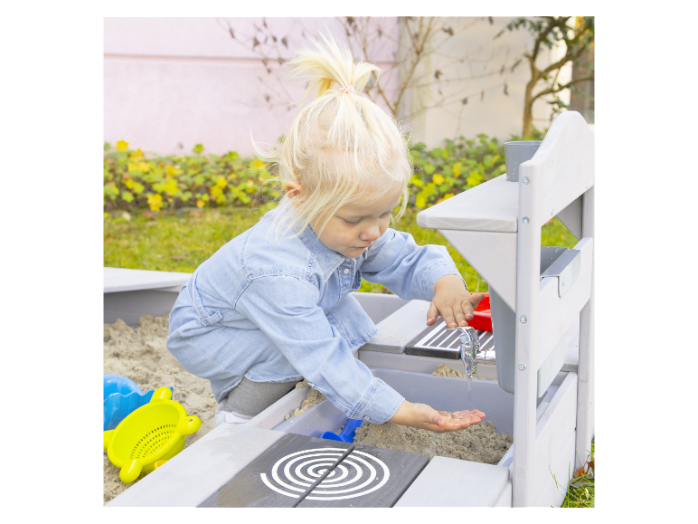 Ein kleines Mädchen spielt in einem Sandkasten mit Wasserhahn und Spielzeug.