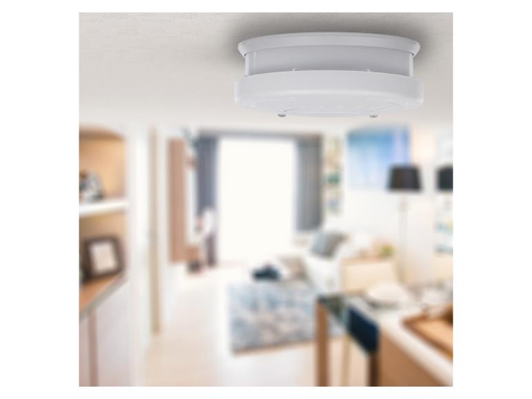Ein weißer Rauchmelder an der Decke in einem unscharfen Wohnzimmer.
