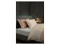 Schlafzimmer mit einem Bett mit grauem Kopfteil, hellgrauem Spannbettlaken, zwei Satin-Kissen in Pfirsich und Creme sowie einem gemusterten Bettbezug. Eine kleine weiße Lampe auf einem Nachttisch beleuchtet die Ecke.