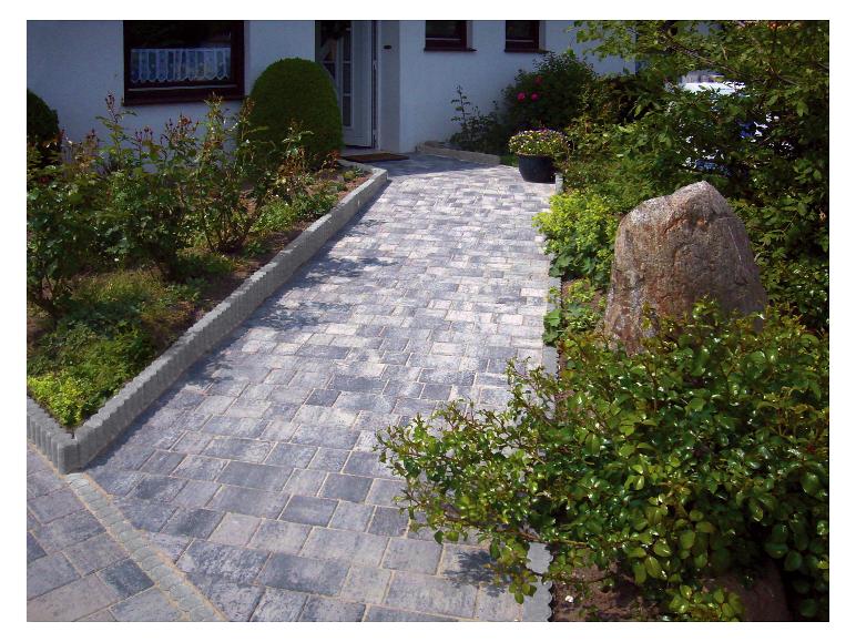 Gepflasterter Weg, umgeben von grünen Büschen und Blumen, führt zu einem Haus.