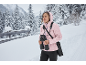 Frau mit Fernglas und rosa Jacke in verschneiter Winterlandschaft.