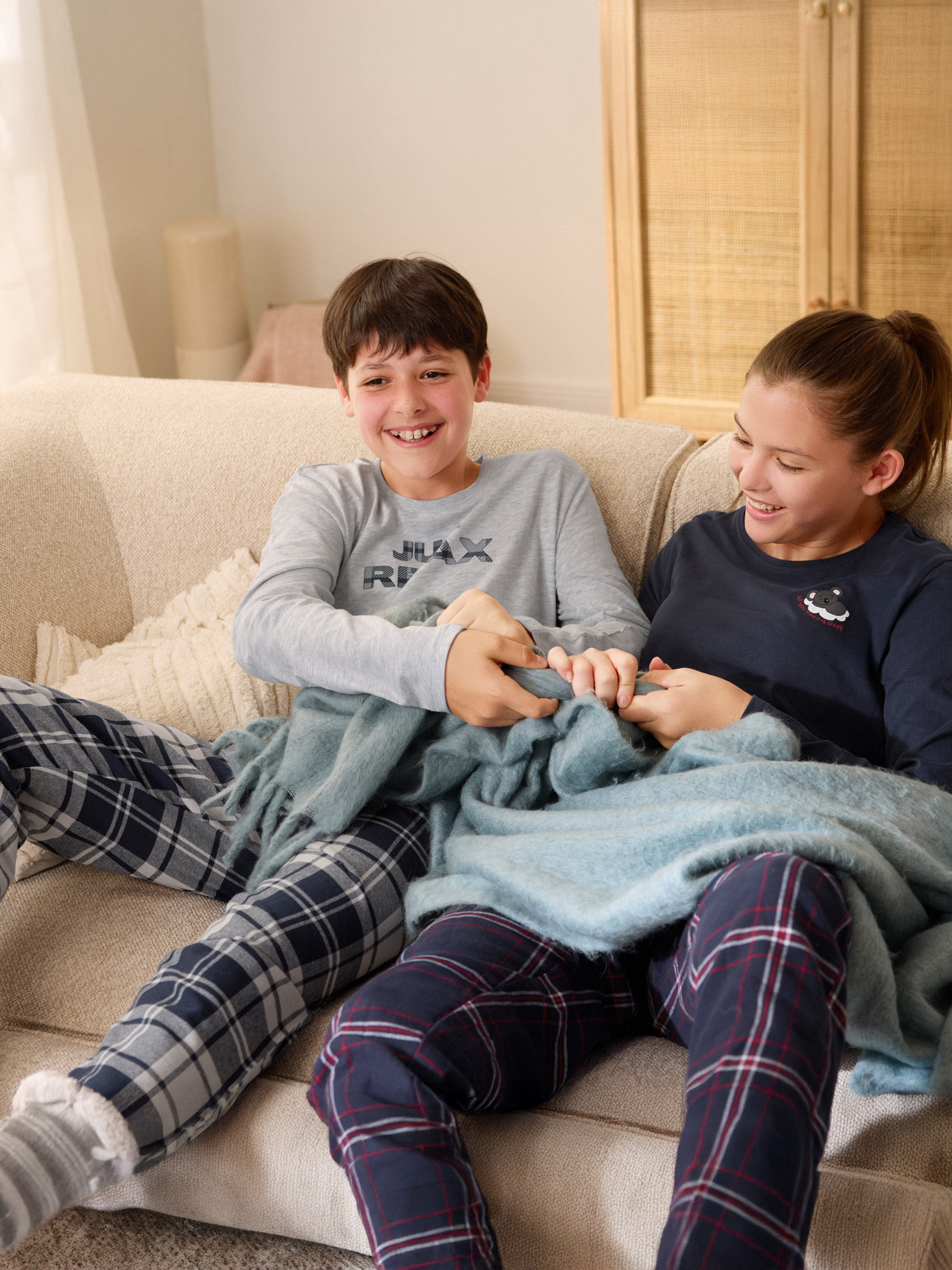 Zwei Teenager in karierten Schlafanzügen mit einer blauen Decke auf einem Sofa.