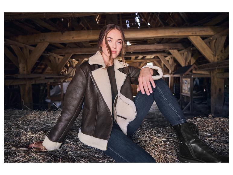Frau in brauner Pilotenjacke und Jeans sitzt auf Heu in einer Scheune.