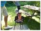 Mann schüttet glühende Kohlen in einen Holzkohlegrill im Garten, Picknicktisch im Hintergrund.