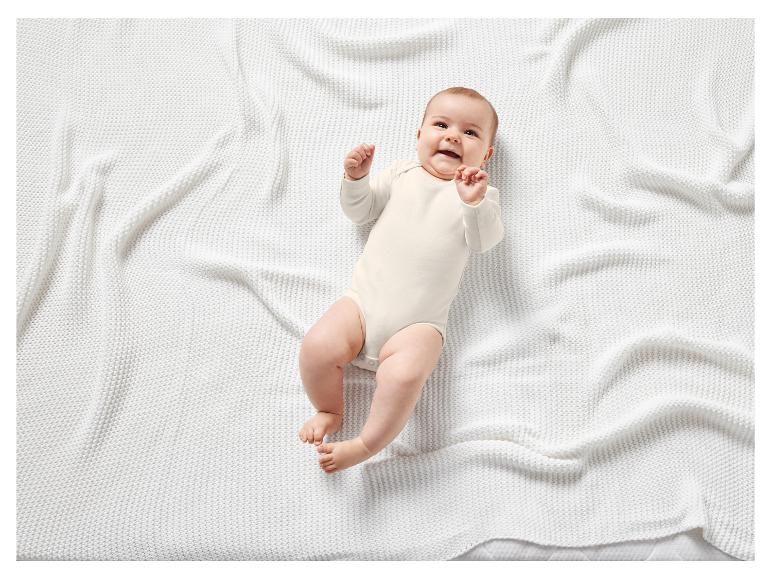 Ein Baby in einem weißen Strampler liegt auf einer weißen Strickdecke.
