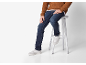 Mann in braunem Strickpullover, blauen Jeans und weißen Sneakern, sitzt auf einem Hocker.