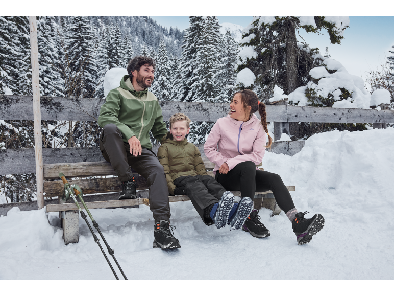 Familie in Winterjacken und Wanderschuhen sitzt auf einer verschneiten Bank und genießt die Winterlandschaft.