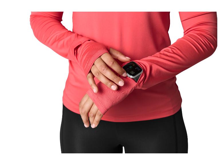 Frau in korallenfarbenem Langarmshirt mit Daumenlöchern, prüft eine Smartwatch am Handgelenk.