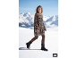 Frau in Leoparden-Strickkleid und schwarzen Stiefeln geht durch den Schnee, Berge im Hintergrund.