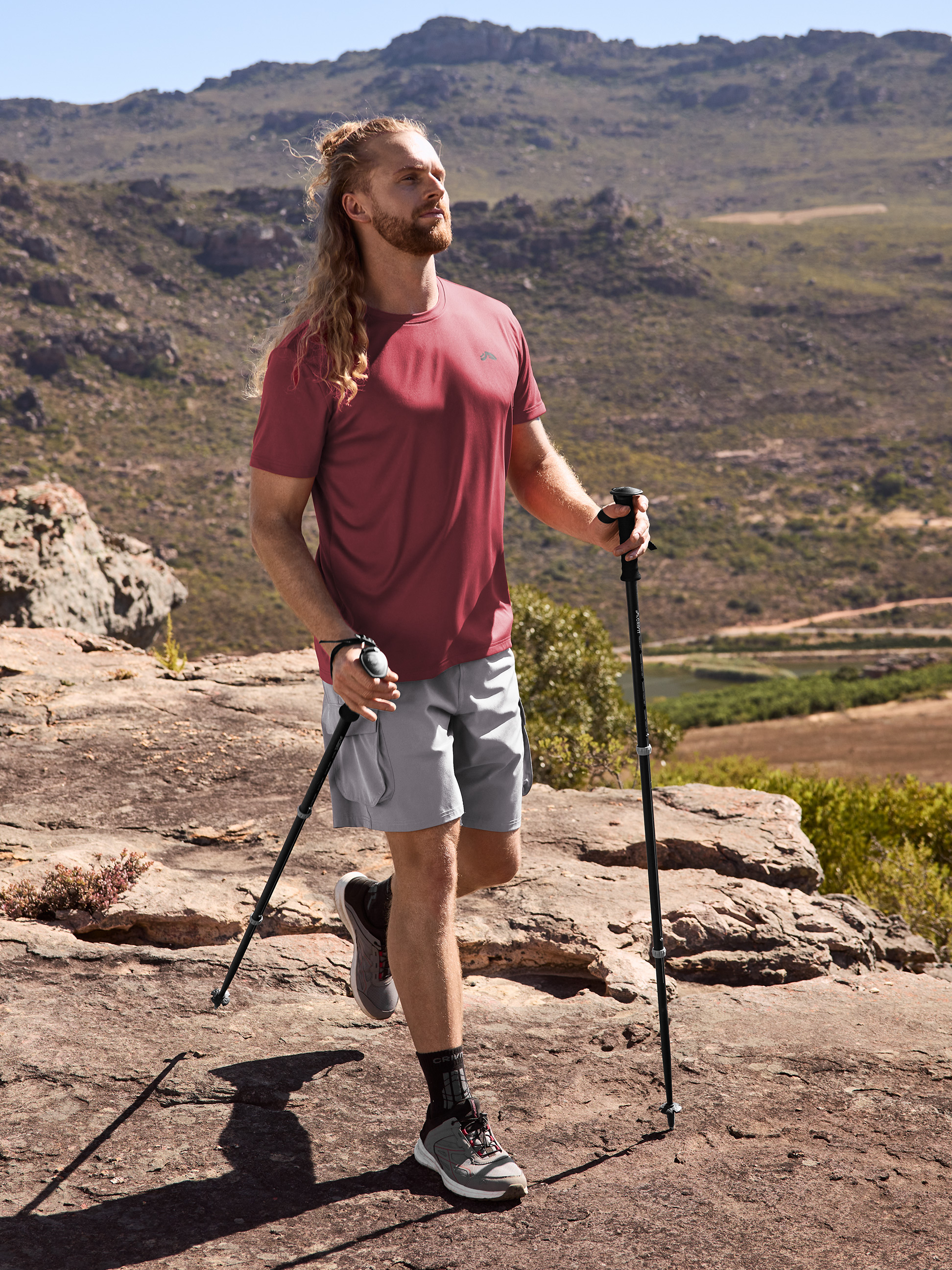 Mann in Wander-T-Shirt und Shorts mit Trekkingstöcken in bergiger Landschaft.