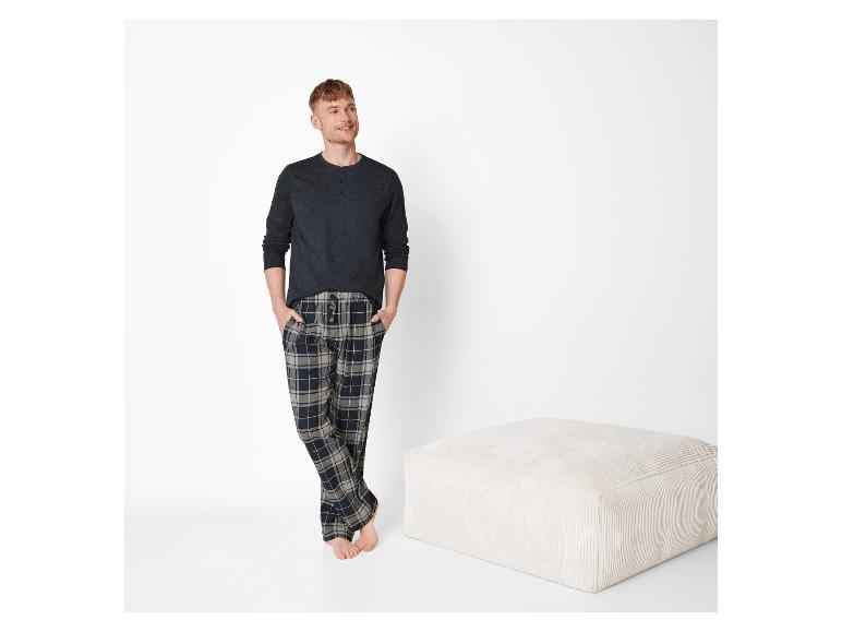 Mann in kariertem Pyjama und grauem Hemd, steht neben einem beigen Hocker.