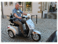 Ein Mann mittleren Alters fährt einen silbernen Dreiradroller auf einer Kopfsteinpflasterstraße.