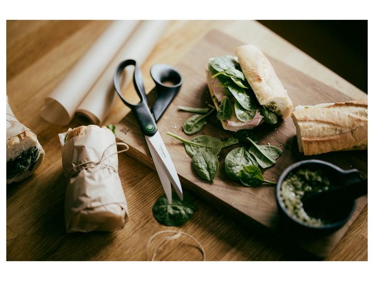 Baguette-Sandwiches mit Spinat und Pesto, Schere und Packpapier auf einem Holztisch.