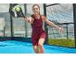 Frau in einem weinroten Sportkleid spielt Padel mit Schläger und Ball.