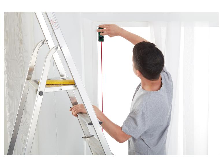 Ein Mann misst ein Fenster mit einem Laser-Entfernungsmesser, während er auf einer Leiter steht.