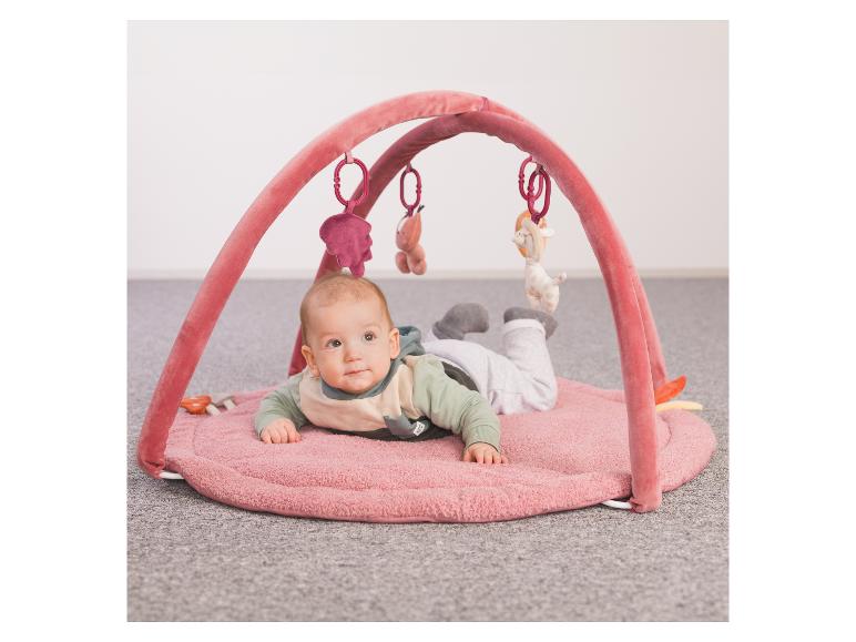 Ein Baby liegt auf dem Bauch auf einer rosa Spielmatte, unter einem Bogen mit hängendem Spielzeug.
