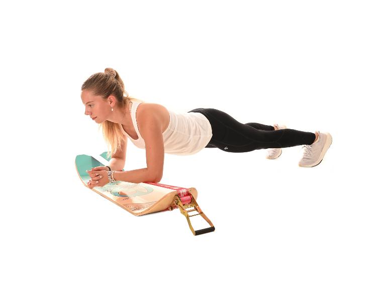 Frau macht Plank auf einem Balance Board mit Widerstandsbändern, trainiert drinnen.