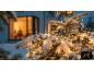 Schneebedeckte Outdoor-Weihnachtsbeleuchtung an einem Baum, mit warm beleuchtetem Fenster im Hintergrund.