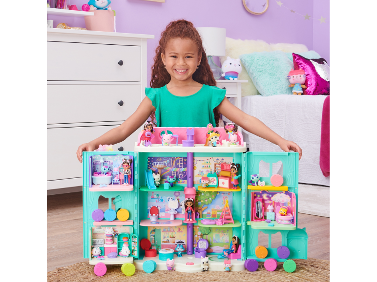Ein lächelndes Mädchen hinter einem bunten, mehrstöckigen Puppenhaus mit Spielfiguren.