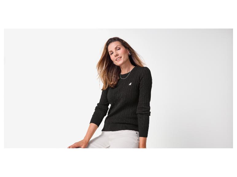 Lächelnde Frau in schwarzem Zopfstrickpullover und heller Hose.