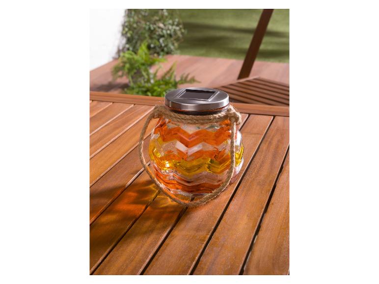 Eine gelbe, orange und weiße Glas-Solarlampe mit einem Seilgriff auf einem Holztisch.