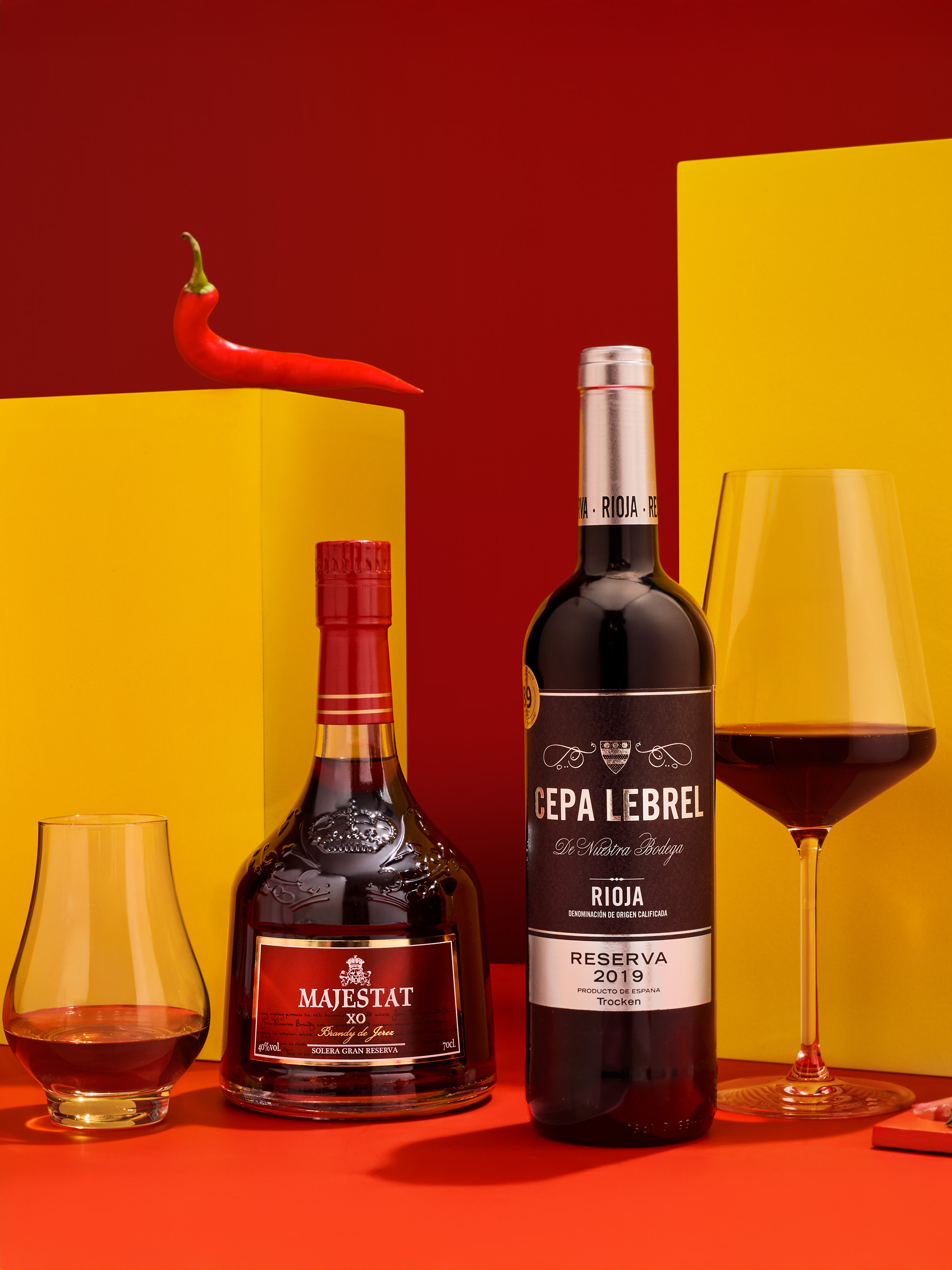 Eine Flasche Rotwein und eine Flasche Brandy mit Gläsern, vor rotem und gelbem Hintergrund.