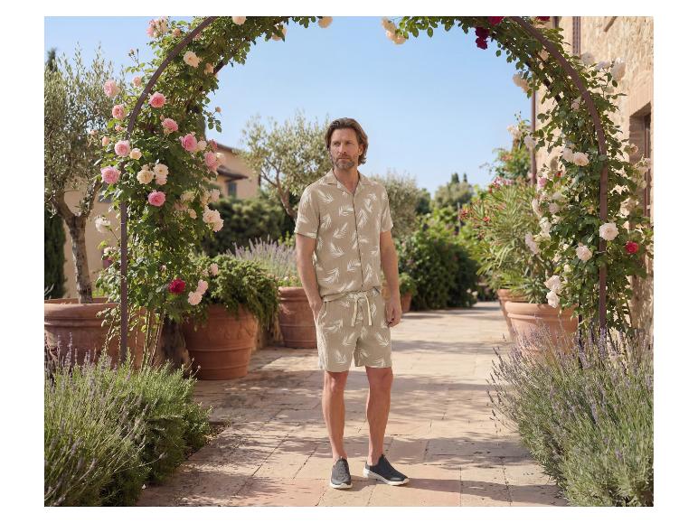 Mann in beigem Hemd und Shorts mit Blattmuster, in einer Gartenumgebung.