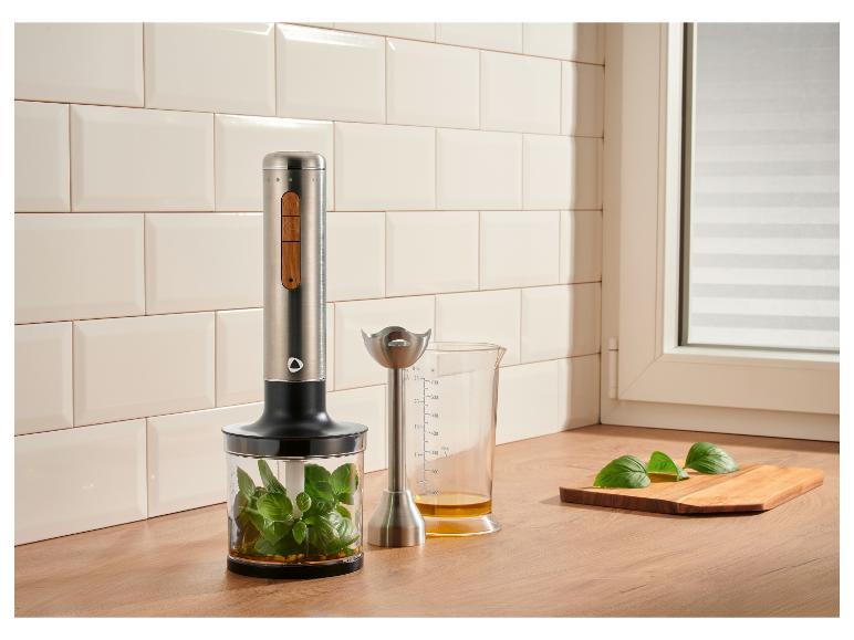 Stabmixer mit Basilikum in Behälter und Messbecher mit Öl auf Holzarbeitsplatte.