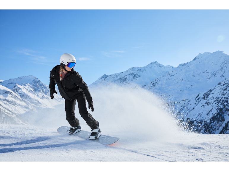Eine Snowboarderin in schwarzer Skijacke und -hose, weißem Helm und blauer Skibrille fährt einen verschneiten Berg hinunter.