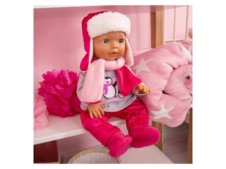 Puppe in rosa Winterkleidung mit Schal und Mütze, sitzt auf einem weißen Regal.